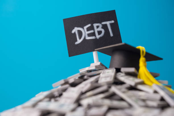istock Mortarboard on heap US dollar banknotes and DEBT label with blue background. 2180921524