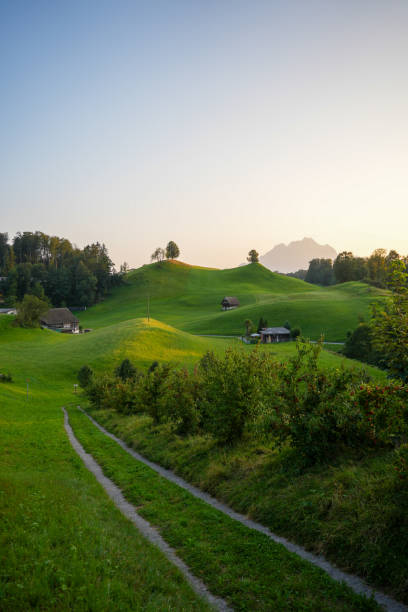 A peaceful rural scene at sunset, featuring rolling green hills dotted with farmhouses and a winding path, with soft evening light casting a warm glow.