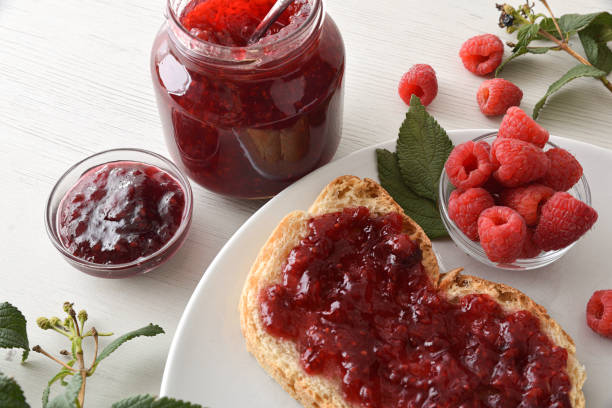 Toast with raspberry jam on white plate with bowl full of berries and jars of jam on wooden bench. Top elevated view.
