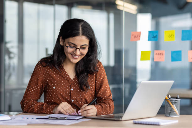 istock Young woman smiling while working at desk in modern office with laptop and notes surrounding her, embodying productivity and focus in professional workspace setting 2180954483