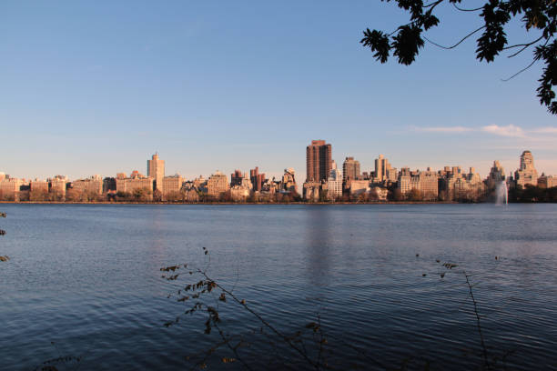 This stunning photograph captures a picturesque view of a city skyline reflected in calm waters, likely taken from a vantage point in a nearby park. The serene lake contrasts beautifully with the towering buildings, which showcase a range of architectural styles. The warm light of the afternoon sun bathes the scene in a golden hue, enhancing the autumn colors and adding to the tranquil atmosphere. This image encapsulates the harmony between urban life and nature, making it an ideal representation of city living, outdoor relaxation, and the beauty of natural landscapes juxtaposed with modern architecture.