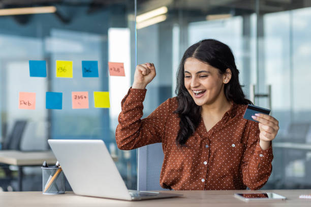 istock Latino businesswoman celebrates successful online transaction while using laptop in modern office 2180955412