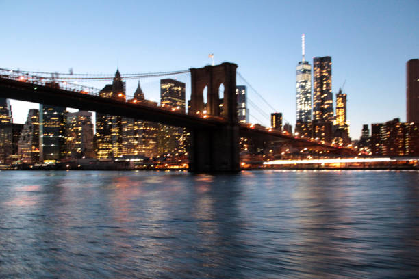 A stunning view of the Brooklyn Bridge at dusk, illuminated against the backdrop of the sparkling New York City skyline. The serene waters of the East River reflect the lights from the bridge and the towering buildings, including the iconic One World Trade Center. The combination of the bridge’s architectural beauty and the glowing cityscape creates a magical and iconic image of New York at twilight. Perfect for themes of cityscapes, urban architecture, and the beauty of New York City’s landmarks.