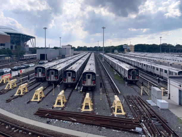 A view of multiple subway trains parked in a vast train yard in New York City. The trains are neatly aligned on multiple tracks, with several more on the adjacent tracks waiting for service or storage. Overhead, a cloudy sky adds a moody atmosphere to the industrial scene. The stadium roof in the background and other city infrastructure frame the busy rail yard, highlighting the essential role of public transit in the city’s daily operations. This image is ideal for themes of transportation, urban infrastructure, and the inner workings of a metropolitan transit system.