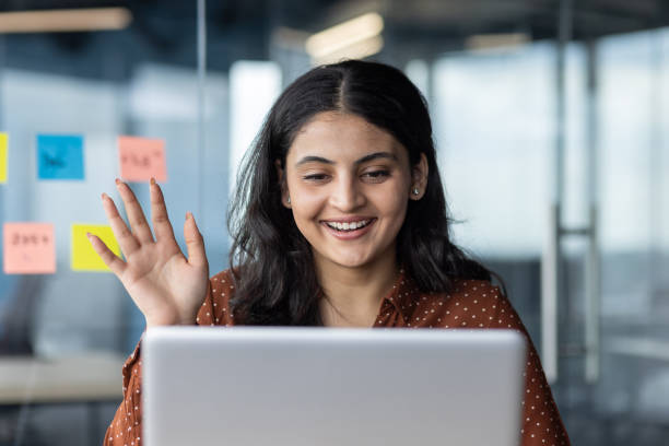 istock Smiling woman engaging in virtual meeting, waving at laptop screen, creating a joyful connection during online conversation in modern office environment with sticky notes in background. 2180956524