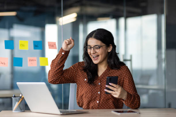 istock Excited woman celebrating success at work using laptop and holding smartphone in modern office environment 2180957904