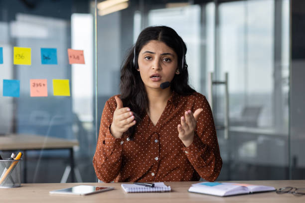 istock Professional woman in office delivering presentation with headset, using hand gestures and focused expression, at work table with notepad and smartphone, emphasizing communication skills 2180958092