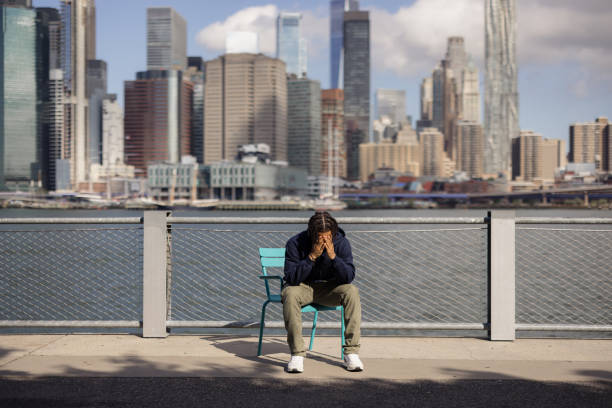 Depressed African American man crying in despair while sitting on a chair by the river in New York city.
