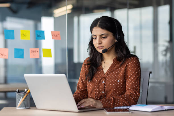 istock Focused woman working at desk using laptop and wearing headset in modern office environment. 2180959252