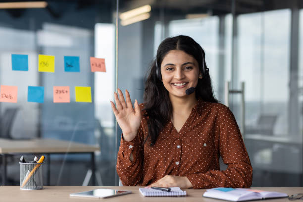 istock Smiling woman with headset waving during online meeting in modern office setting with colorful sticky notes on glass wall 2180959418