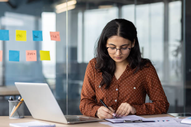 istock Focused woman working on paperwork in modern office with laptop and sticky notes on glass wall 2180961148