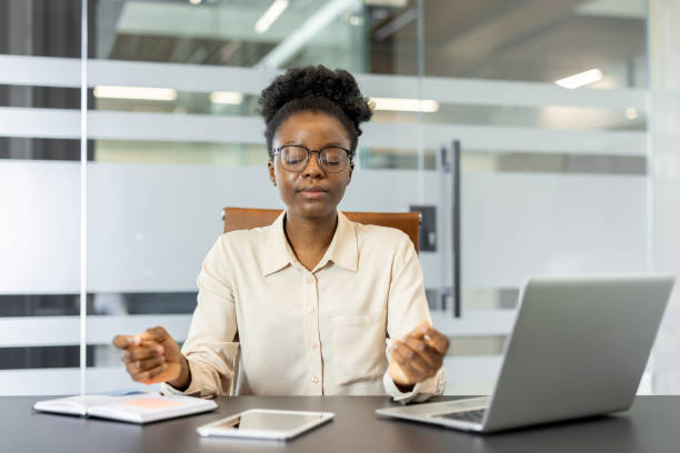 Calm African American businesswoman practicing mindfulness during office break at desk with eyes closed, embracing relaxation for improved focus and productivity in workplace