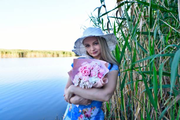 Beautiful woman in the evening in summer on the lake with a bouquet of sunflowers and a bouquet of roses.