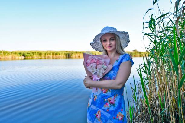 Beautiful woman in the evening in summer on the lake with a bouquet of sunflowers and a bouquet of roses.