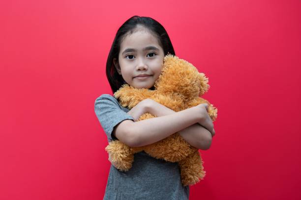 Little Asian girl hug teddy bear against red background.