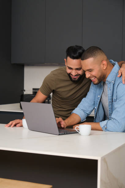istock Happy gay couple using laptop and smiling while having coffee in kitchen 2180990780