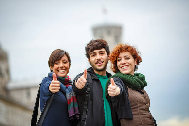 Happy students gesturing thumbs up in front of the leaning tower of pisa stock photo