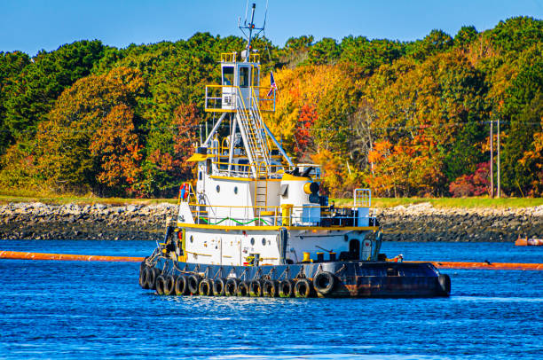 With a backdrop of colorful autumn trees along the bank of the Cape Cod Canal, a yellow and white tugboat assists other boats in the area laying dredging pipes in the waterway.