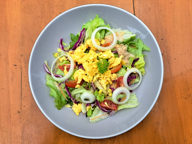 Delicious homemade gourmet healthy chicken salad bowl, with Spanish onion, cherry tomato, lemon and olive oil, on a garlic and lettuce green salad, served in a rustic handmade ceramic bowl, with seasoning and herbs on a white rustic natural wooden kitchen or restaurant table, representing a healthy lifestyle, wellbeing and body care, top view with a copy space