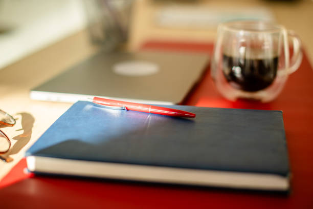 A workspace featuring a blue notebook, red pen, and clear glass cup on a red mat, with a blurred background of a laptop and other office accessories.