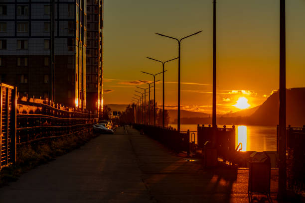 Golden sunset over a peaceful waterfront pathway with silhouetted streetlights and distant hills