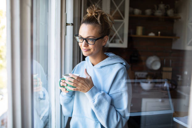 istock Young woman having a cup of coffee while looking through a window at home 2181030672