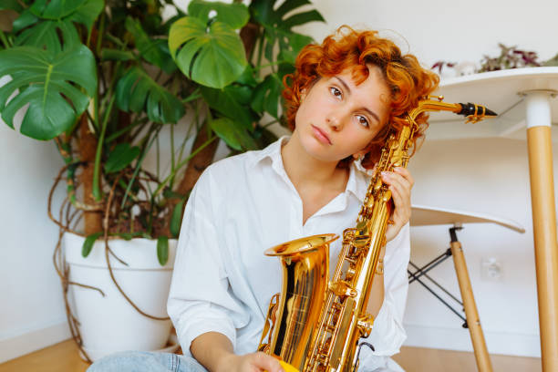 portrait of young attractive red-haired woman with wind musical instrument saxophone in hands sitting on floor in room