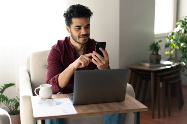istock Happy young man using phone while working with laptop at home 2181048333