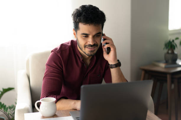 istock Happy young man making phone call and using laptop at home 2181048481
