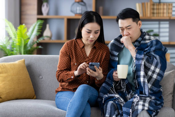 istock Concerned Asian couple using phone for online doctor's consultation at home, supporting each other during illness with cozy setup including blanket and hot drink for comfort 2181049252