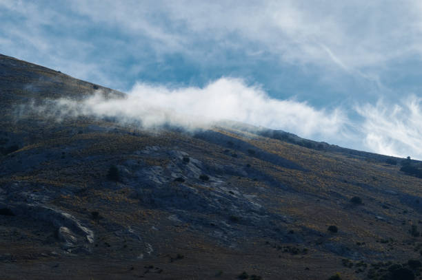 A stunning view of a rugged mountain landscape with swirling clouds and a clear blue sky. The rocky terrain is bathed in natural sunlight, creating a serene and atmospheric scene.