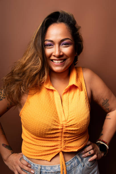 Studio portrait of a cheerful young latin woman with tattoos, smiling and posing with hands on hips against a brown background