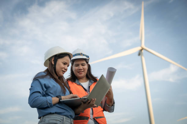 istock two engineers are looking at a laptop while standing next to a wind turbine. They are wearing hard hats and safety vests, indicating that they are working in a construction or maintenance setting 2181068788