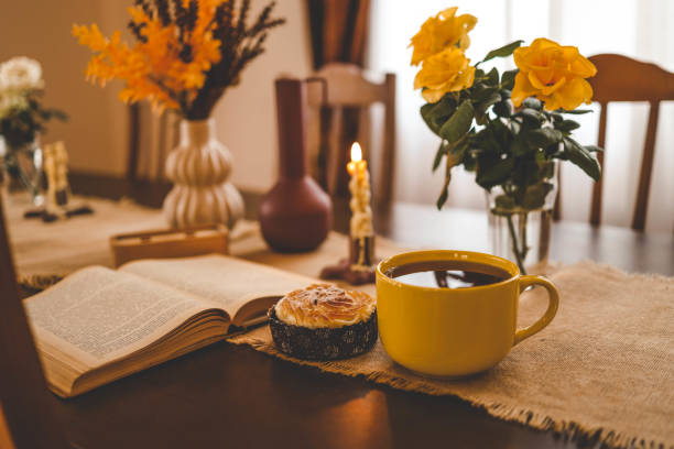 Cozy dining table with a warm autumnal atmosphere featuring a yellow cup, an open book, a muffin, and decorative vases with yellow flowers. The scene evokes a sense of relaxation and comfort.
