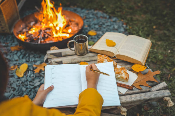 istock Person writing in a notebook next to a bonfire during autumn. Outdoor cozy setting with fallen leaves, snacks, hot drink, and the warmth of the fire, capturing the essence of a peaceful autumn day 2181080417