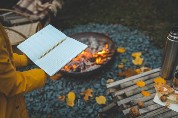 istock Person writing in a notebook next to a bonfire during autumn. Outdoor cozy setting with fallen leaves, snacks, hot drink, and the warmth of the fire, capturing the essence of a peaceful autumn day 2181080420