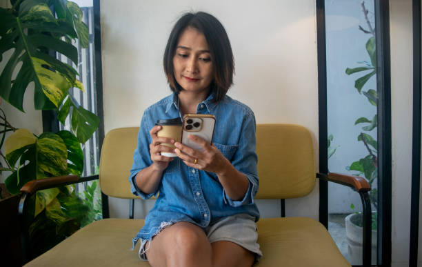 istock Asian woman sitting on sofa hand holding coffee cup and using mobile smart phone shopping online relaxing holiday concepts 2181107166