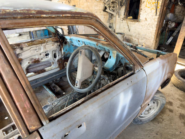 An old, white and red Soviet-era liftback car is abandoned in tall grass near a dilapidated building in the countryside
