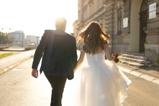 Rear view shot of cheerful newlyweds holding hands and walking in the city on sunny day