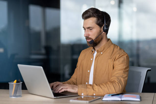 istock Man in brown shirt working on a laptop with headset in modern office setting, focused and engaged in his task. 2181137371