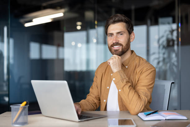 istock Young professional smiling confidently while working on laptop in modern office interior with smartphone and stationery creating positive workspace vibe 2181138002