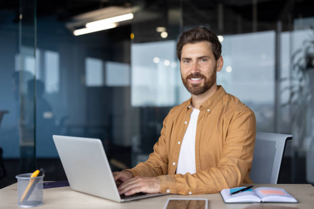 istock Confident professional man sitting at desk with laptop and documents in modern office environment, wearing casual attire, showcasing work efficiency and productivity, looking at camera 2181138361