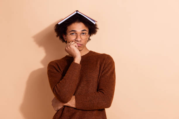 istock Thoughtful young student with book on head in casual sweater pondering on beige background 2181140438