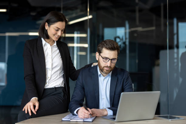 istock Professional colleagues interacting in a modern office setting, with a female employee standing beside a seated male worker at a desk, highlighting workplace dynamics and teamwork. 2181144185