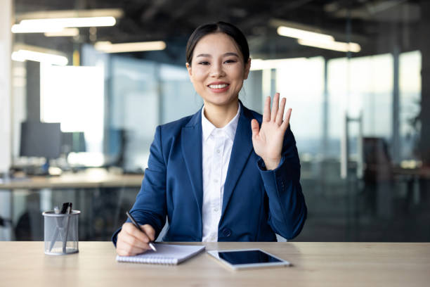 istock Asian businesswoman waving hello in modern office while taking notes and using digital tablet during virtual meeting 2181146712
