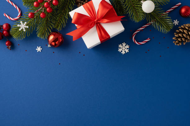 istock Christmas themed image featuring a wrapped gift with red ribbon, pine branches, ornaments, and candy canes on a blue background 2181152750