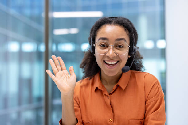 istock Young woman working in a modern office environment using headset for customer support, smiling and waving to camera during online video call or remote meeting 2181154698