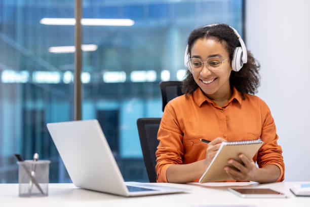 istock Young woman enjoying learning at workplace with laptop and notepad. Smiling while wearing headphones in bright modern office. 2181161341