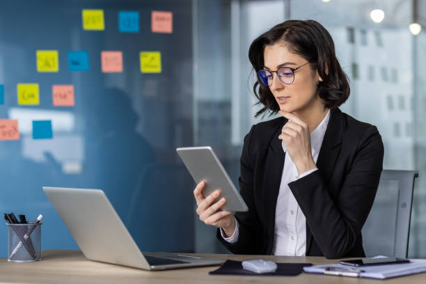 istock Professional woman in business attire using tablet while working in modern office with laptop and organized sticky notes on glass wall 2181174944