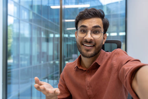 istock Smiling man in glasses making a video call using phone in modern office space looking at camera 2181175419